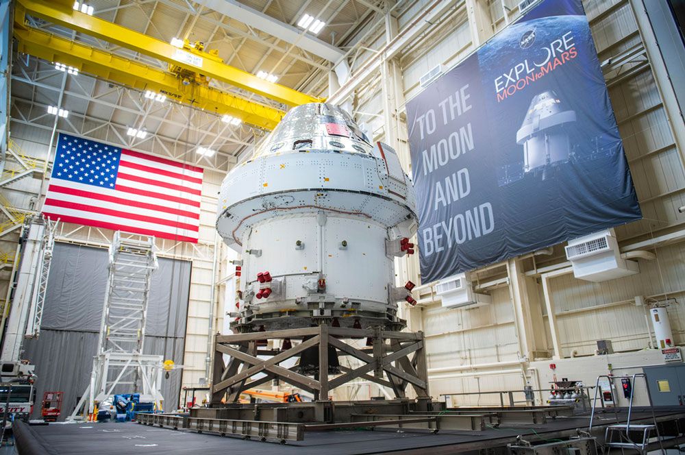 Navicella Orion all’interno di una grande struttura con la bandiera americana su una parete e un cartellone con la scritta “To the Moon and Beyond” (Verso la luna e oltre) sull’altra.