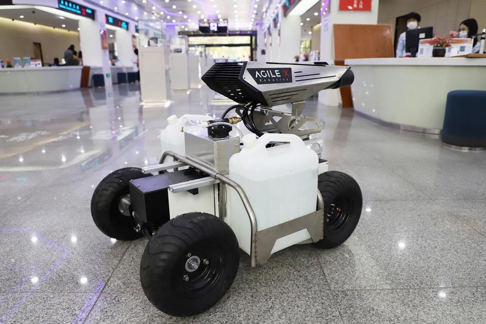 A disinfecting robot carrying two tanks of liquid disinfectant inside a commercial space. It has four wheels and a spray attachment.